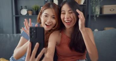 年轻的亚洲女人开心地笑着放松，在家里<strong>客厅</strong>里使用智能手机视频通话。快乐的室友女士与朋友和<strong>家人</strong>的视频会议，家庭妇女的生活方式概念.